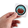 Male hand holding small round Keep Tassie Wild sticker with Tasmanian waratah (telopea truncata)