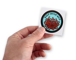 Male hand holding small round Keep Tassie Wild sticker with Tasmanian waratah (telopea truncata)